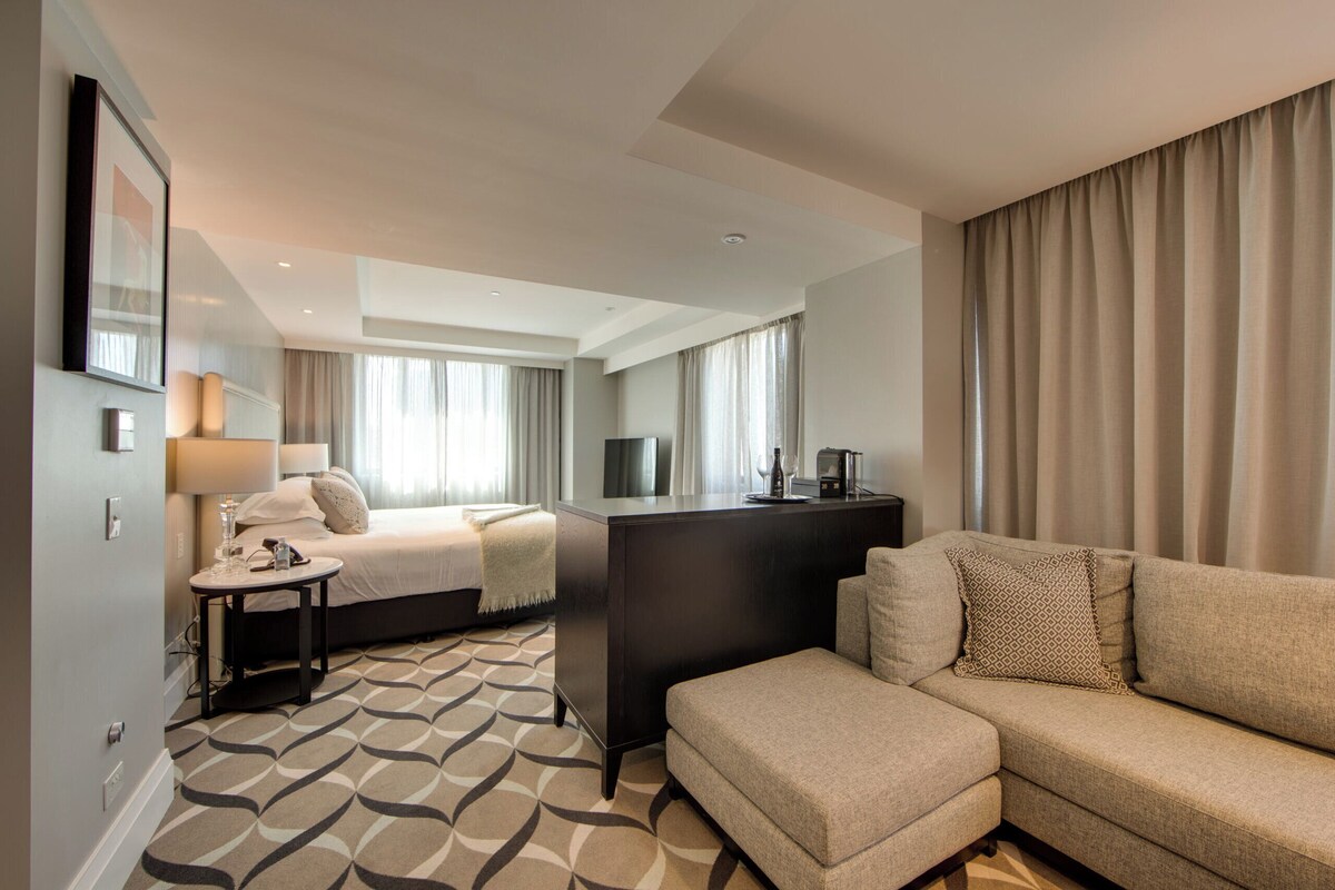 Une chambre d’hôtel moderne, équipée d’un canapé, d’un lit, d’une petite table avec une lampe et d’un téléviseur posé sur un meuble.