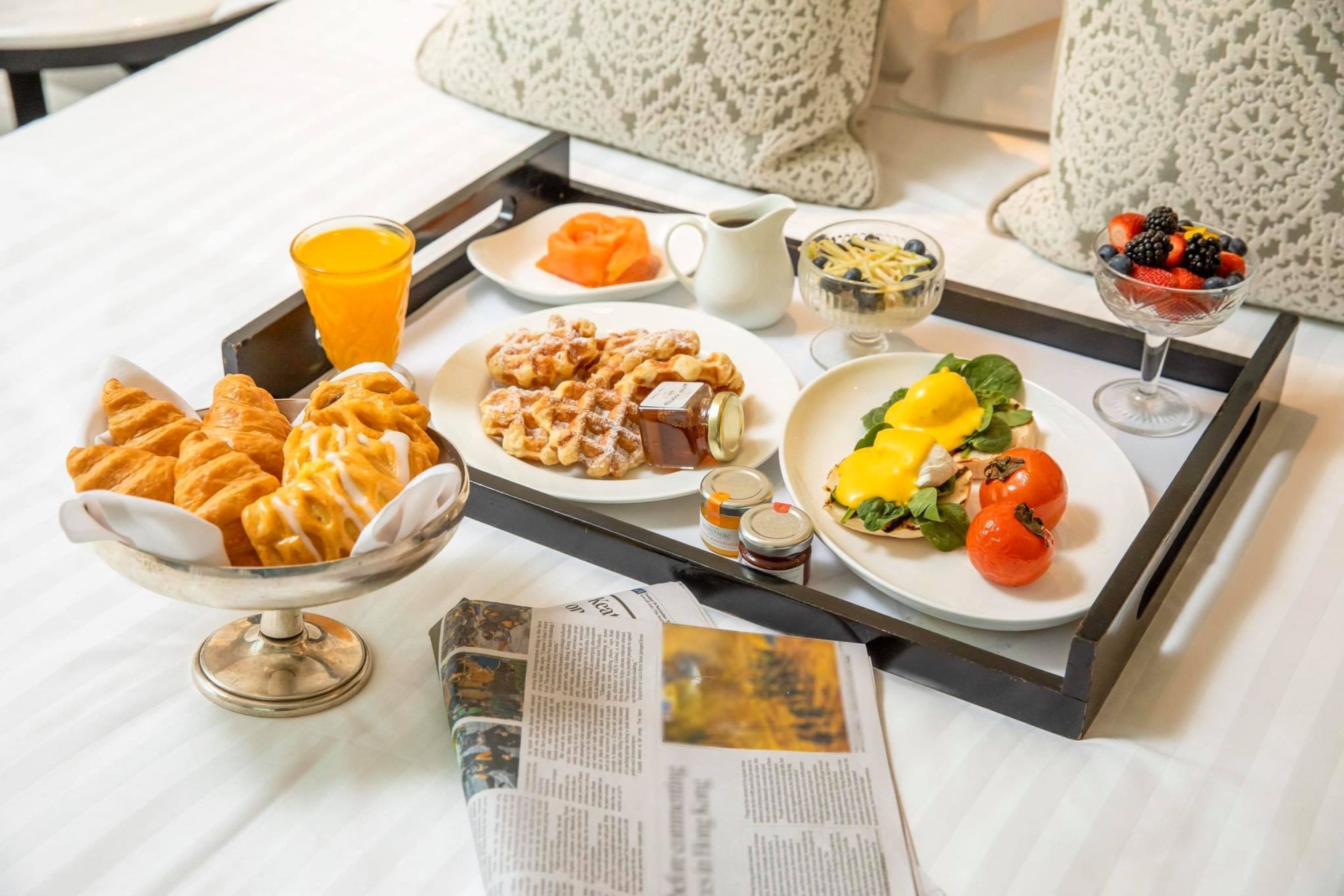 Una bandeja de desayuno con pasteles, fruta y jugo sobre una cama, junto a un periódico.