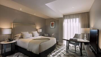Habitación de hotel con una cama grande, mesitas de noche con lámparas, un escritorio con silla, televisión y un ventanal con cortinas.