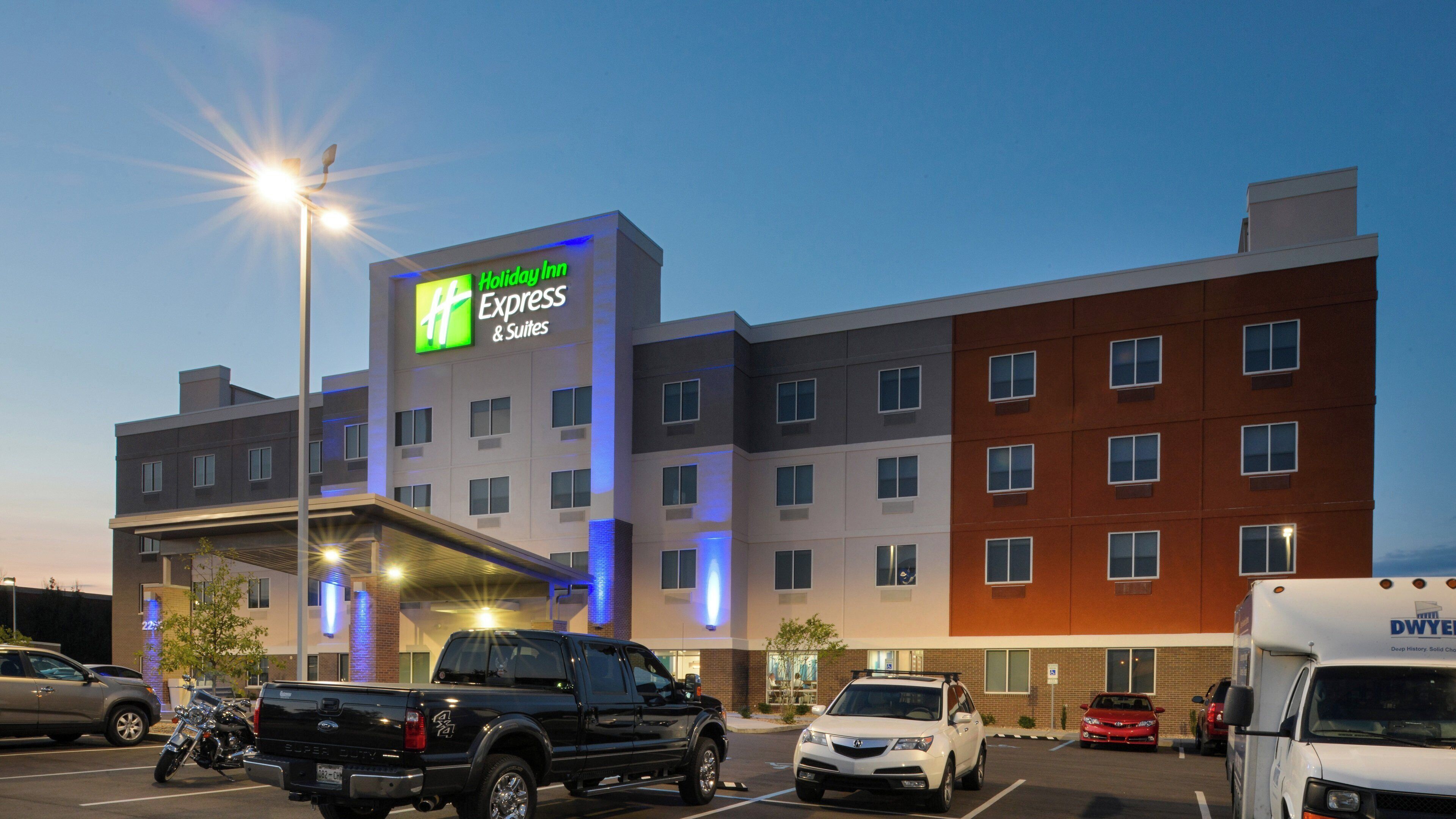 A Holiday Inn Express & Suites hotel with a modern design, multiple floors, and a parking lot with various vehicles.
