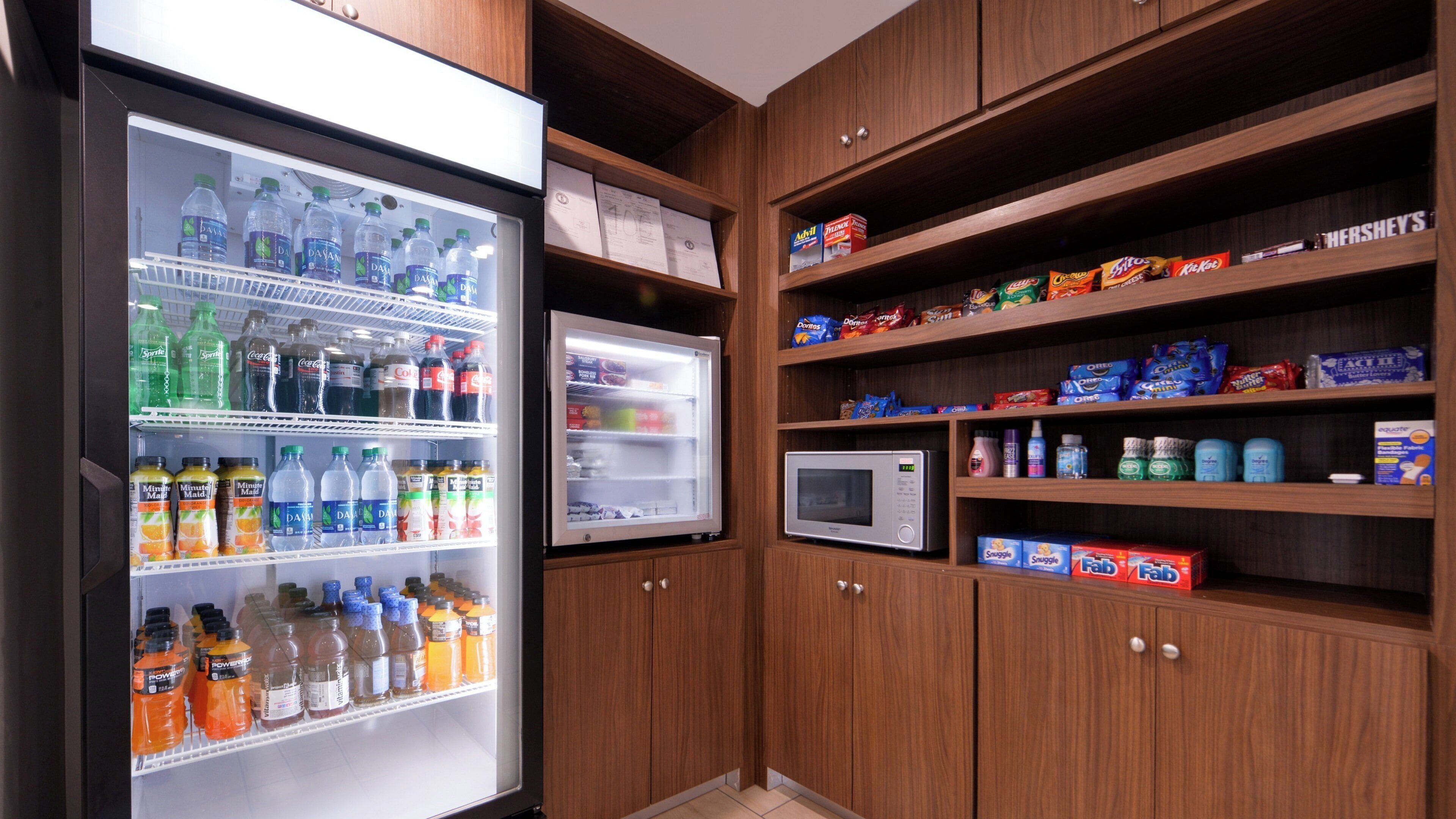 Una kitchenette con un refrigerador lleno de bebidas, un microondas y estantes con aperitivos y artículos de tocador.