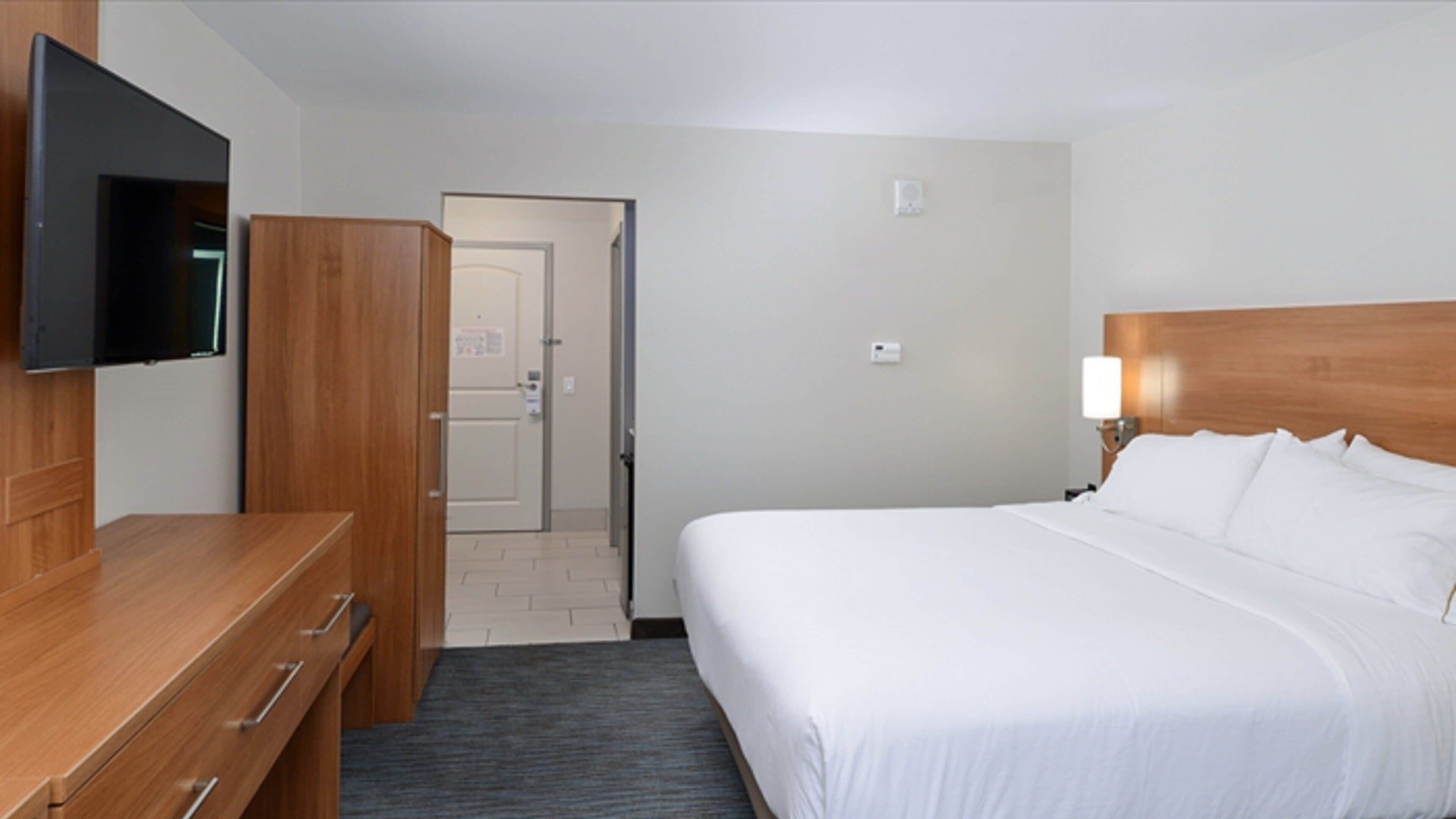 Ein Hotelzimmer mit einem Bett, einem an der Wand befestigten Fernseher, einer Holzkommode und einer Tür zu einem weiteren Raum.