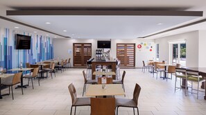 A modern dining area with multiple tables and chairs, a flat-screen TV mounted on the wall, and a view of the outdoors through large windows.