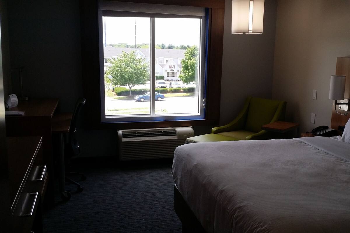 Una habitación de hotel con cama, escritorio, silla, ventana con vista a edificios y coches, y una lámpara de pared.