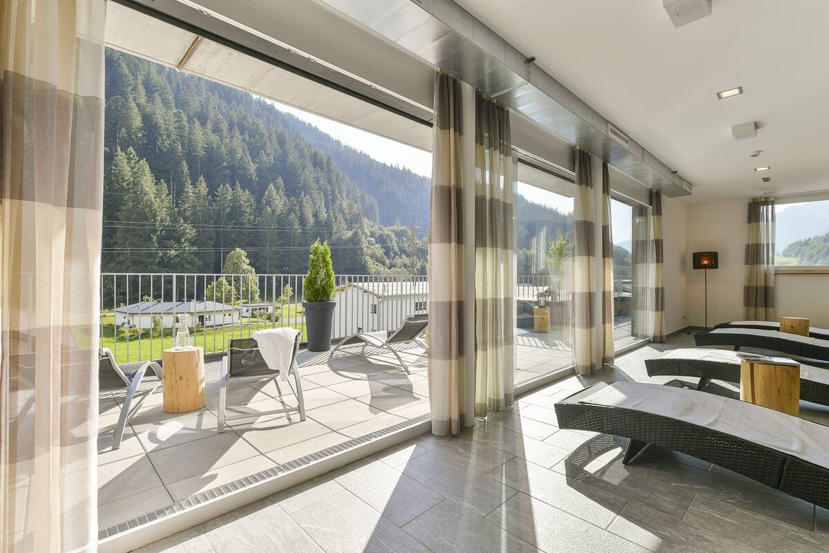 Un balcon avec coin salon extérieur, une vue sur une montagne boisée et un intérieur moderne avec de grandes fenêtres et des rideaux.