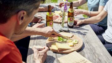 Un grupo de personas alrededor de una mesa de madera con una tabla de quesos, botellas de cerveza y una flor.