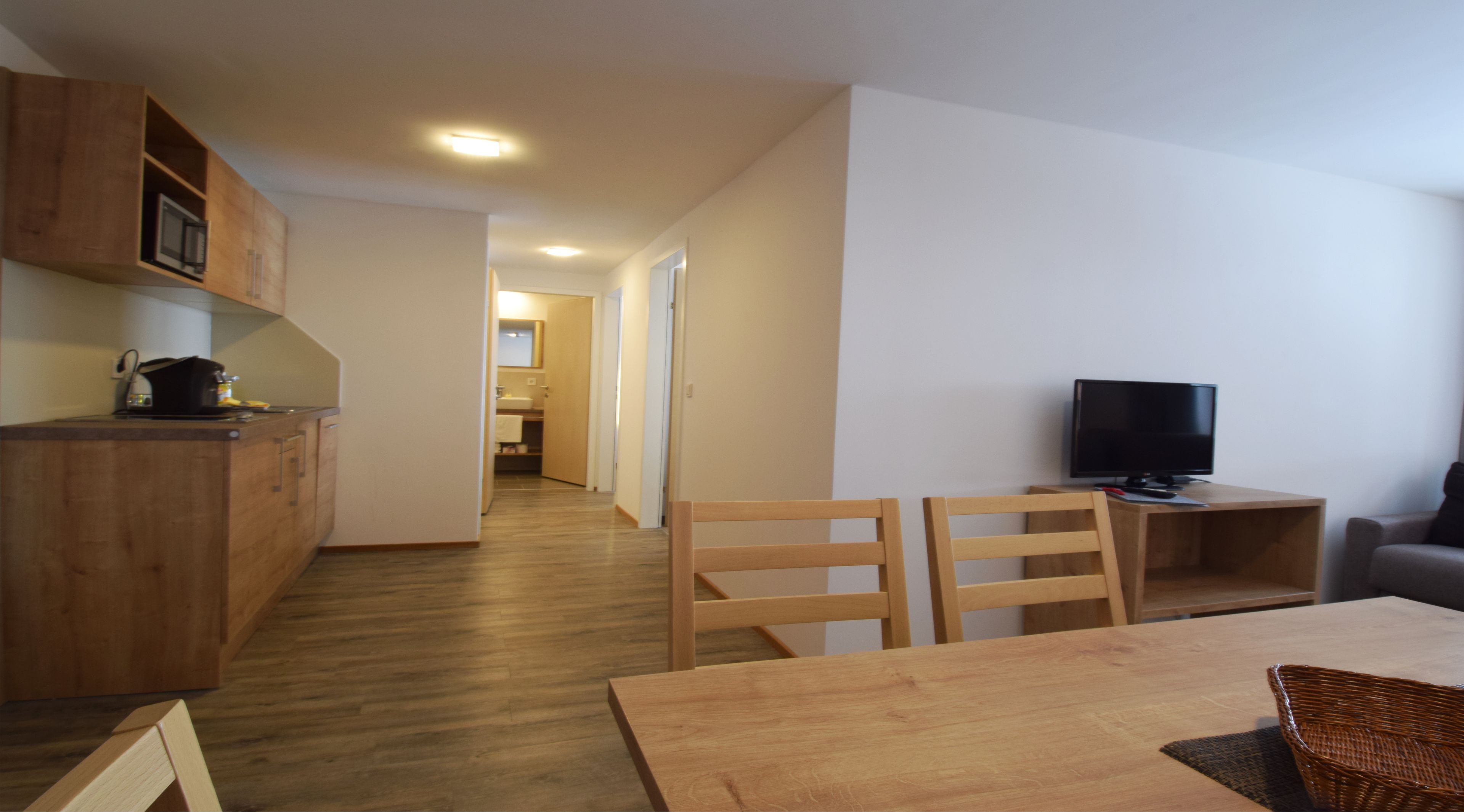 Une cuisine moderne avec des meubles en bois, un coin repas comprenant une table et des chaises, un téléviseur et un couloir menant à une salle de bain.