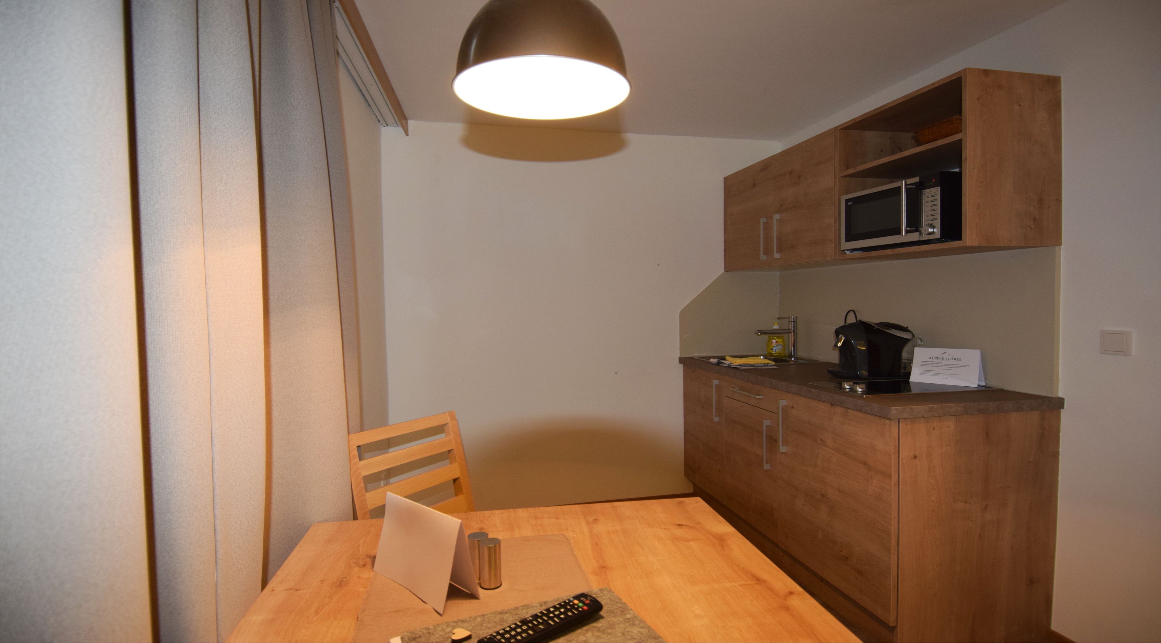 A compact kitchen with wooden cabinets, a microwave, and a coffee maker. There is a wooden dining table with a chair and a remote control on it.