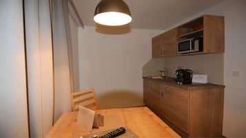 A compact kitchen with wooden cabinets, a microwave, and a coffee maker. There is a wooden dining table with a chair and a remote control on it.