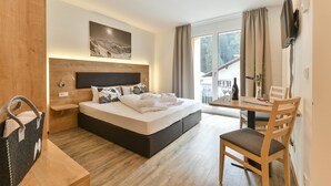A modern hotel room with a large bed, a wooden headboard, a small desk with a chair, a television, and a window with curtains. - Alpine Lodge Klösterle am Arlberg (Klösterle am Arlberg)