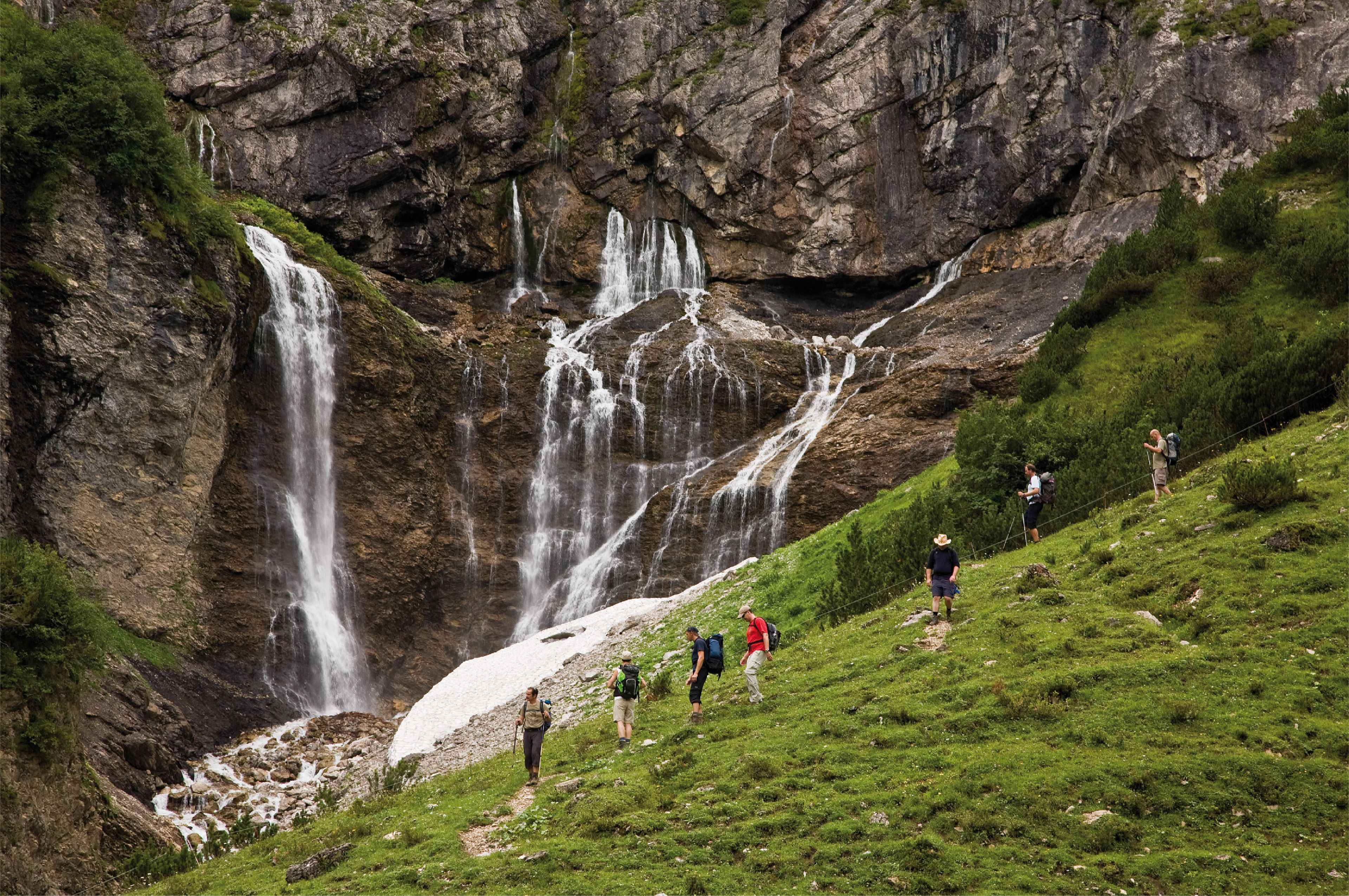 Un grupo de excursionistas en una ladera cubierta de hierba, con una cascada que desciende por un risco rocoso.