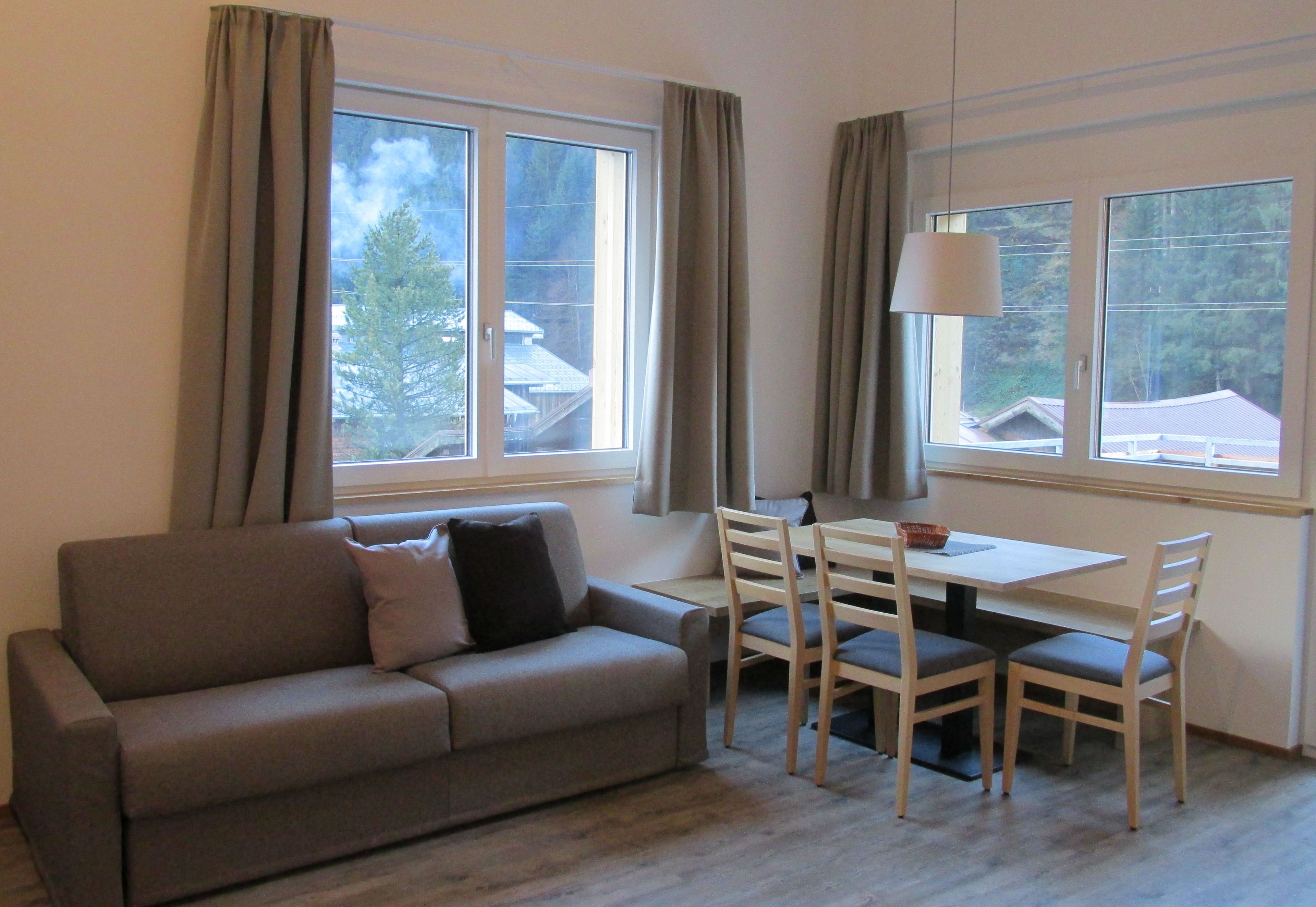 Una habitación con sofá, mesa y sillas, ventanas amplias con cortinas y vistas a edificios.