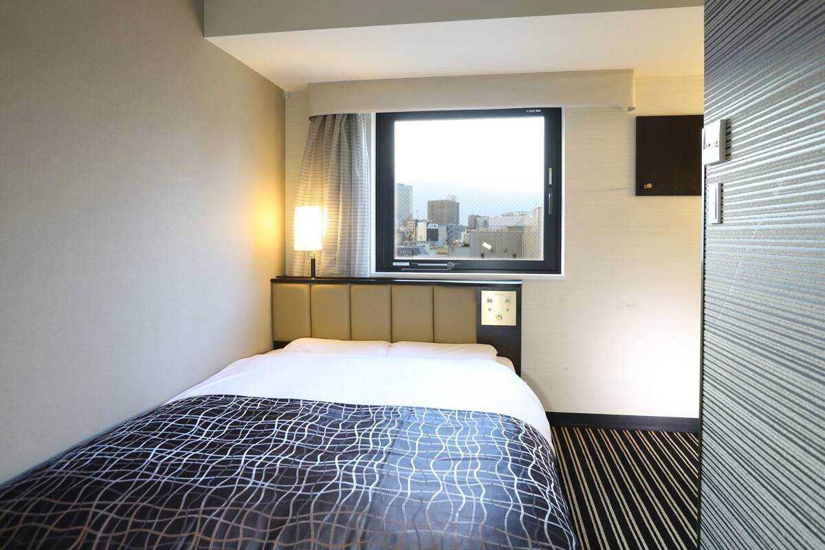 Een hotelkamer met een bed, een raam met uitzicht op de stad, een wandlamp en een bedieningspaneel.