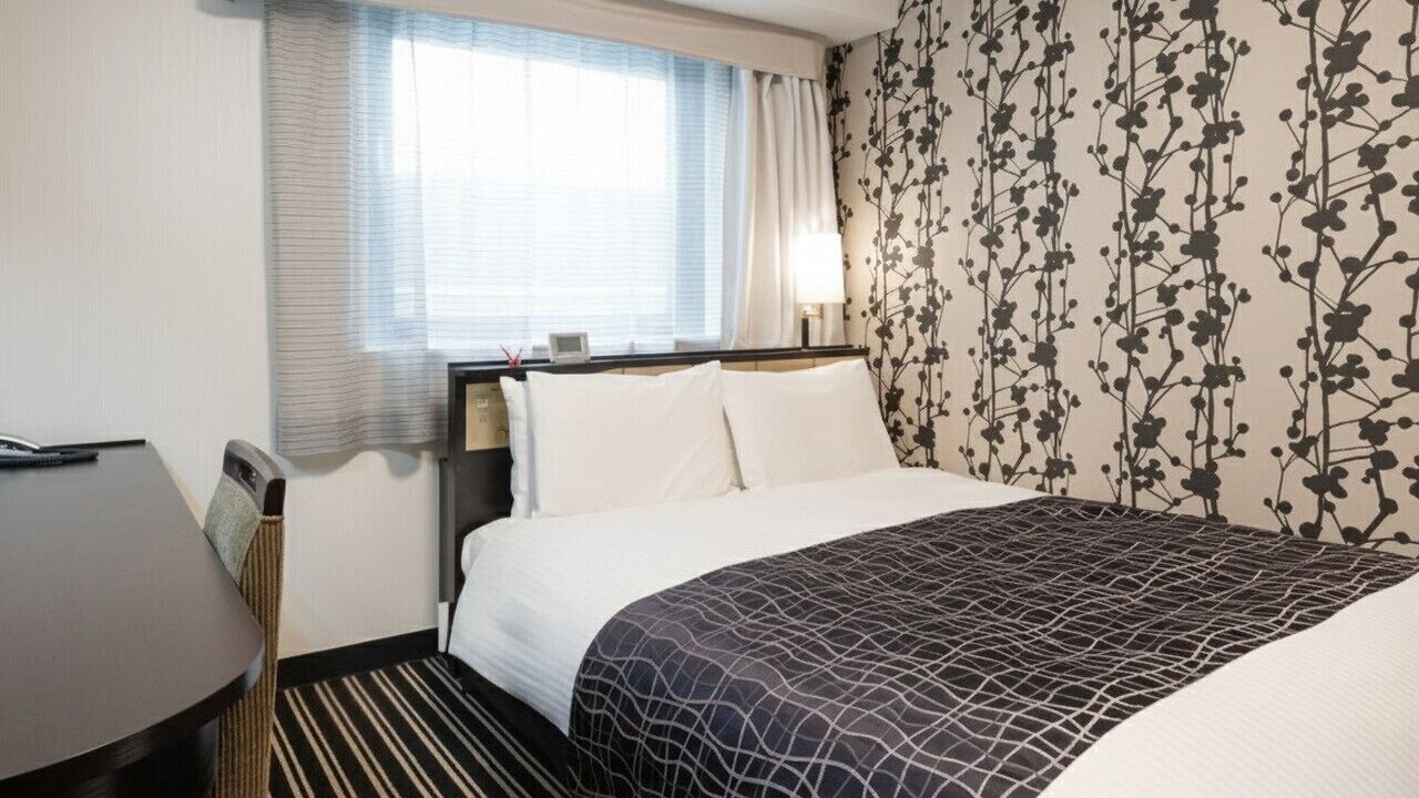 Een hotelkamer met een bed, nachtkastje, lamp, raam met gordijnen en behang met bloemenmotief.