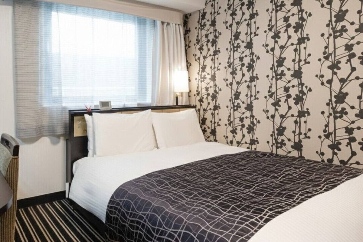 Een hotelkamer met een bed, nachtkastje, lamp, raam met gordijnen en behang met bloemenmotief.