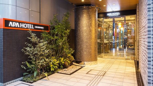 L’extérieur de l’hôtel APA présente une entrée moderne, une grande colonne métallique et un chemin dallé menant à l’entrée.