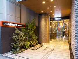 Exterior del APA HOTEL con una entrada moderna, un gran pilar metálico y un camino empedrado que conduce a la entrada.
