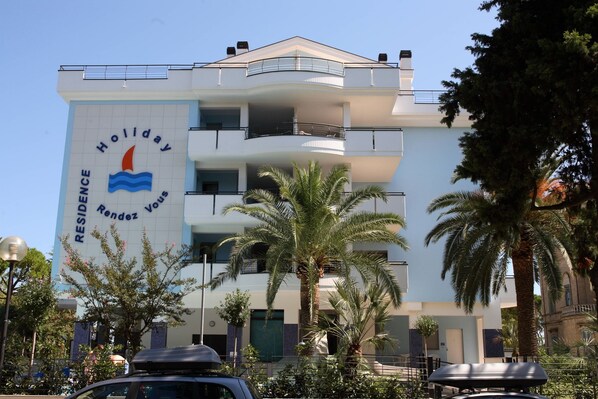 A multi-story building with balconies, palm trees, and a sign reading "Holiday Residence Voulou".