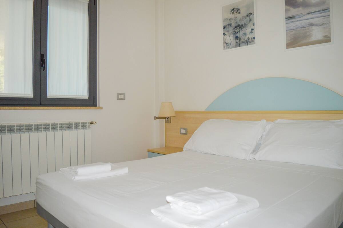 A neatly made bed with white linens and towels, a wooden headboard, a bedside table with a lamp, and a window with blinds.