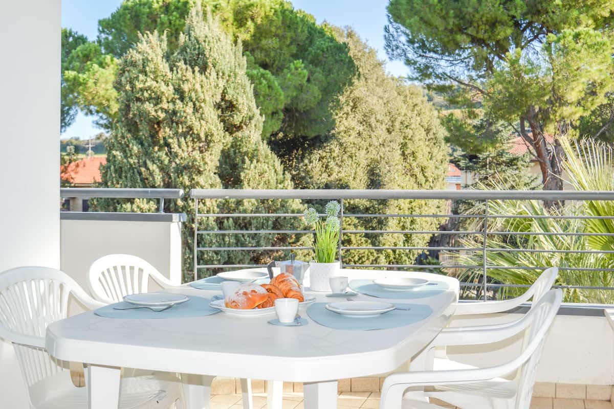 A white table set with plates, cups, and a pastry, surrounded by white chairs, on a balcony with a railing and trees in the background.