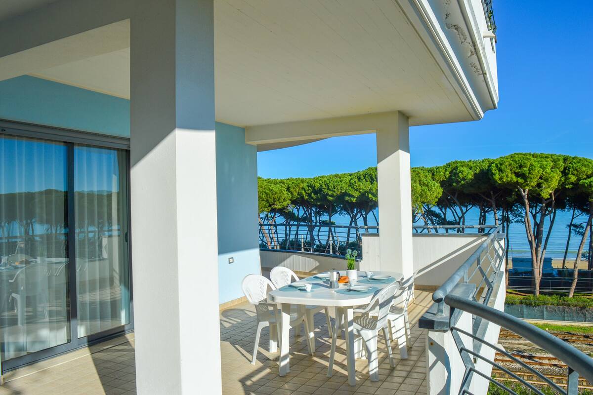 A balcony with a table and chairs, overlooking a coastal area with trees and a clear sky.