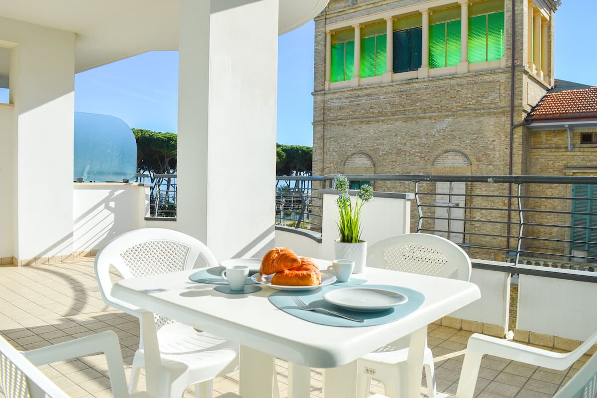 A balcony with a table set for breakfast, including croissants and a cup, overlooking a historic building with green shutters.