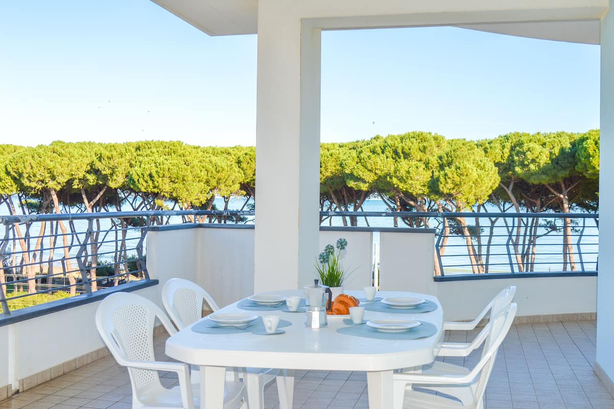 A balcony with a white table set for dining, overlooking a vineyard and a body of water.