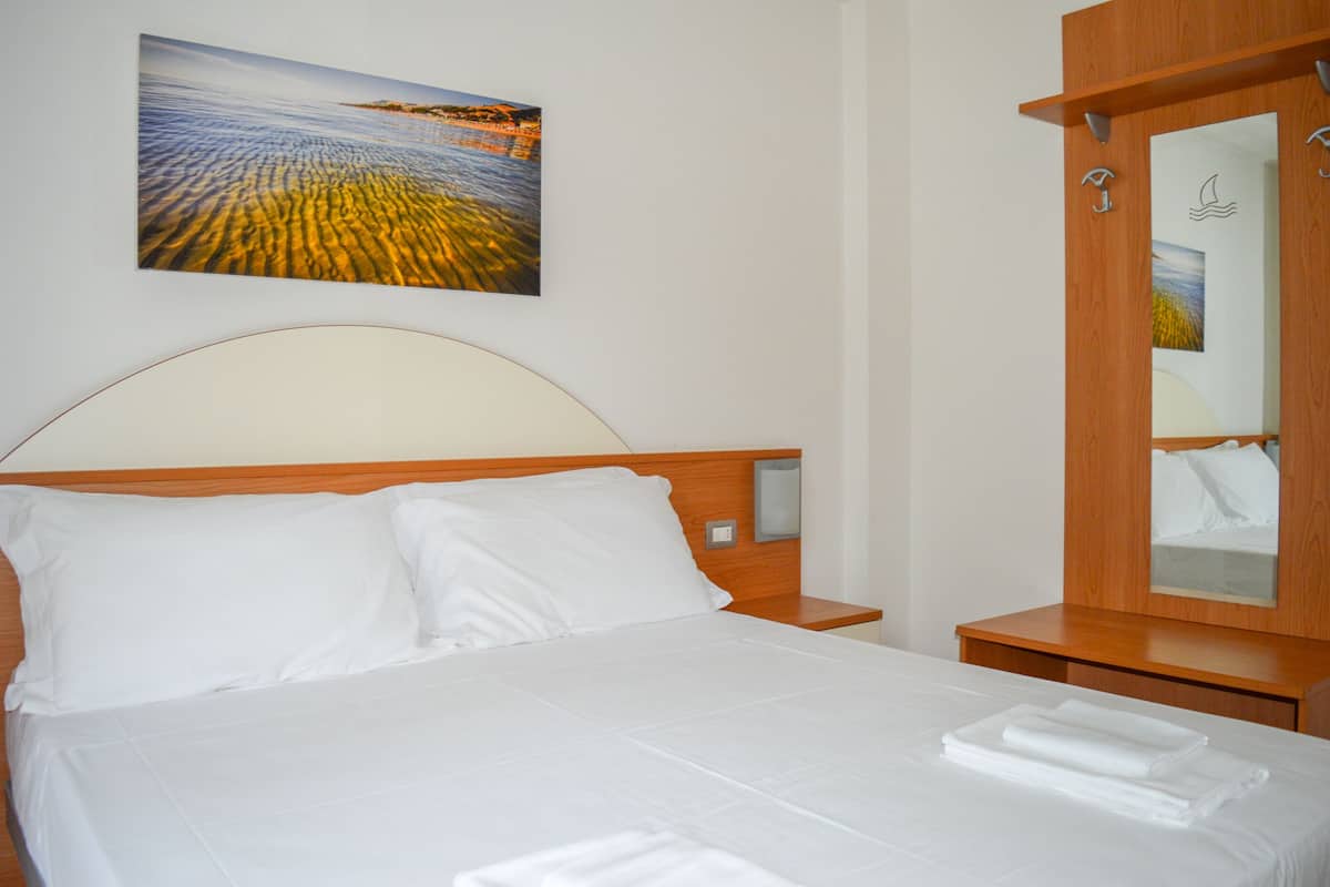 A neatly made bed with white linens and pillows, a wooden headboard, a bedside table, and a wall-mounted painting of a landscape.