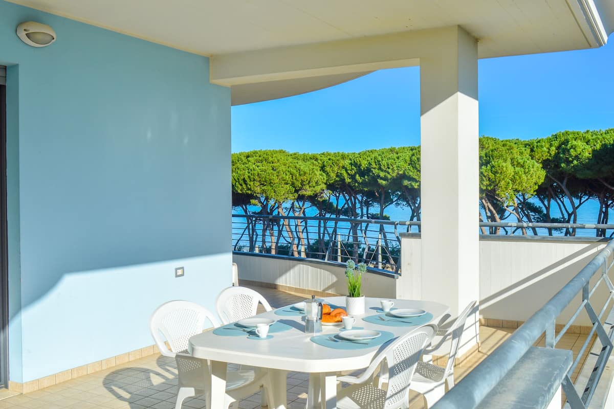 A balcony with a table set for two, white chairs, and a view of the sea and trees.