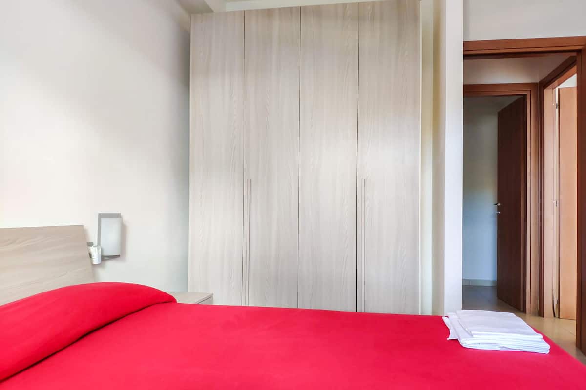 A room with a bed, a wooden wardrobe, a nightstand, and a wall-mounted light.