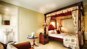 A bedroom with a four-poster bed, a bedside table with lamps, a small table with a pitcher, and a chair.