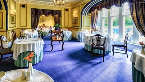 Uma sala de jantar formal com várias mesas preparadas para o serviço, grandes janelas com cortinas e um candelabro.