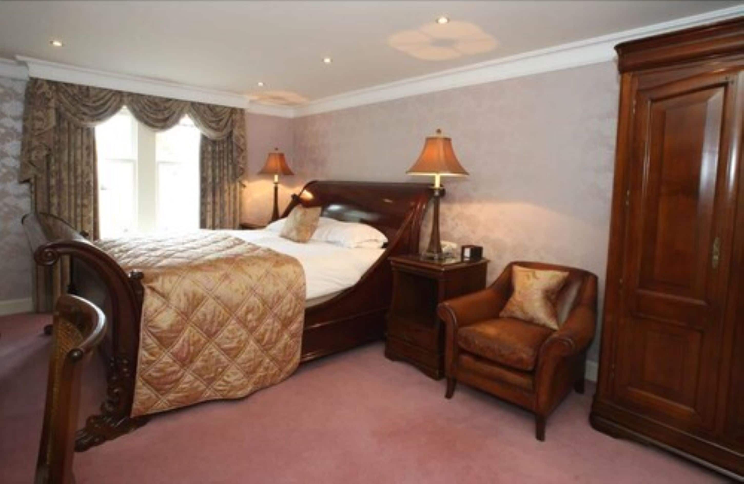 A bedroom with a large bed, two bedside lamps, a wooden wardrobe, and a leather armchair.