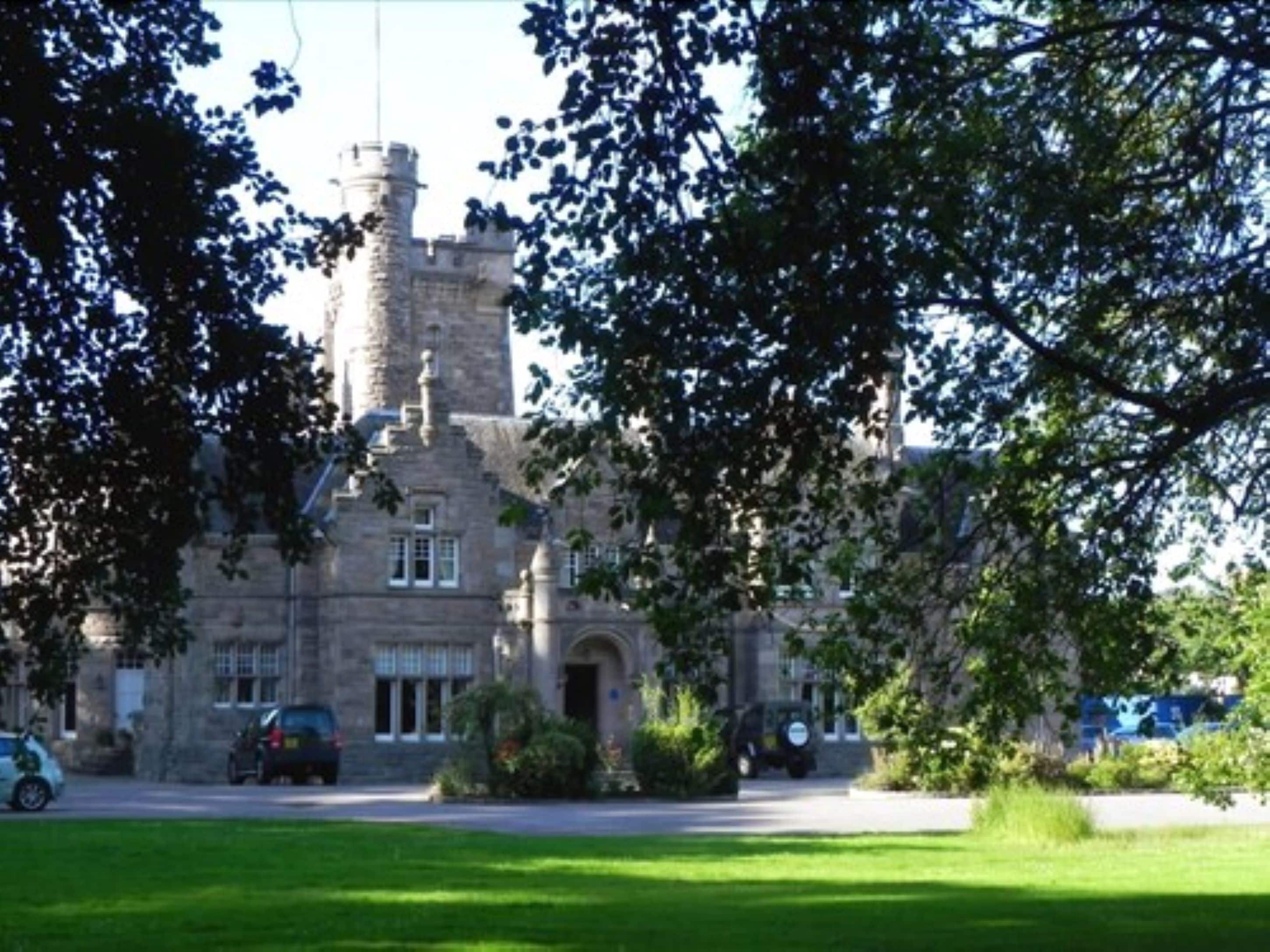 A castle-like building with a tower, surrounded by greenery and parked cars.