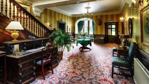 Uma sala imponente com uma escadaria, um candelabro, uma mesa com toalha verde e uma planta grande.