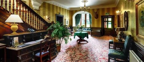 A grand room with a staircase, a chandelier, a table with a green tablecloth, and a large plant.