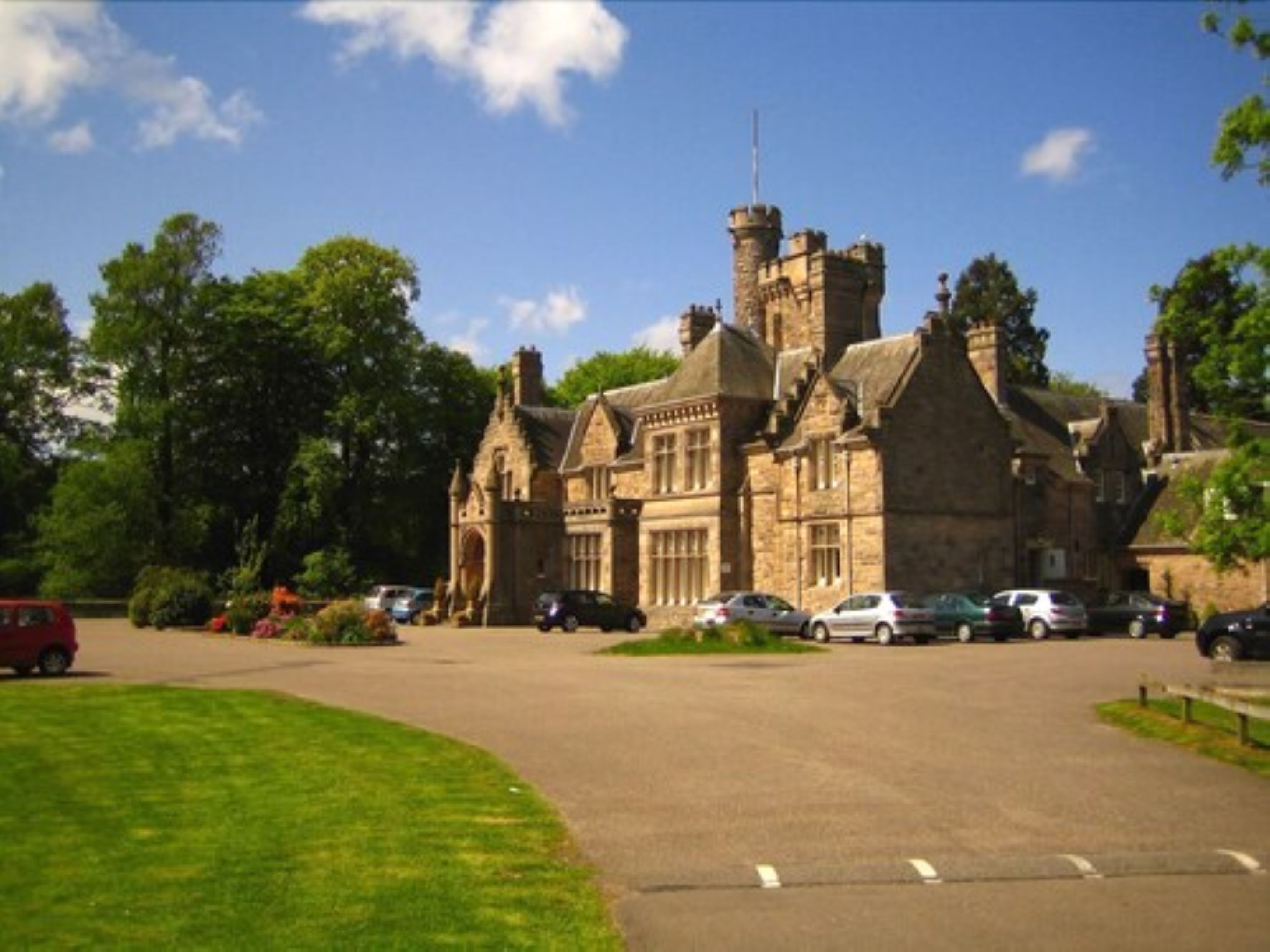 A large stone building with a prominent tower and arched windows, surrounded by greenery and parked cars.