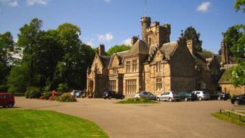 A large stone building with a prominent tower and arched windows, surrounded by greenery and parked cars.