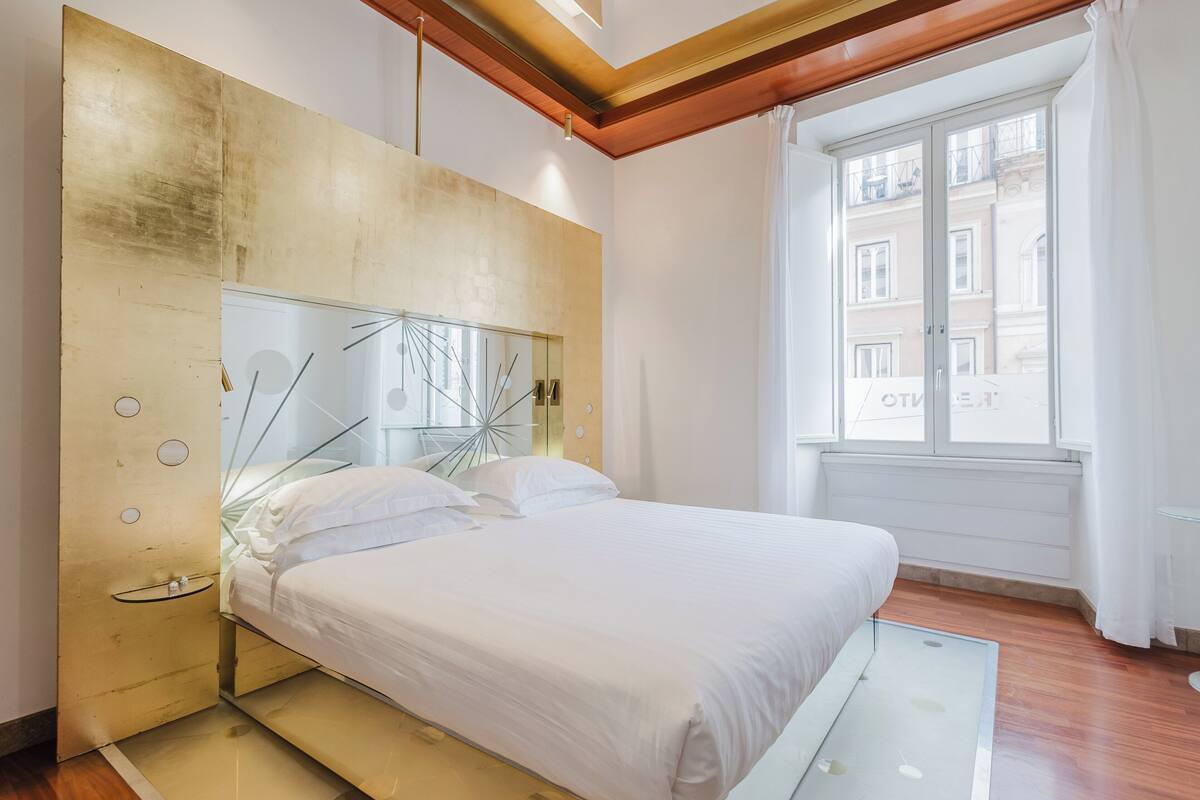 A modern bedroom with a large bed, a glass headboard, and a view of a building through a window.