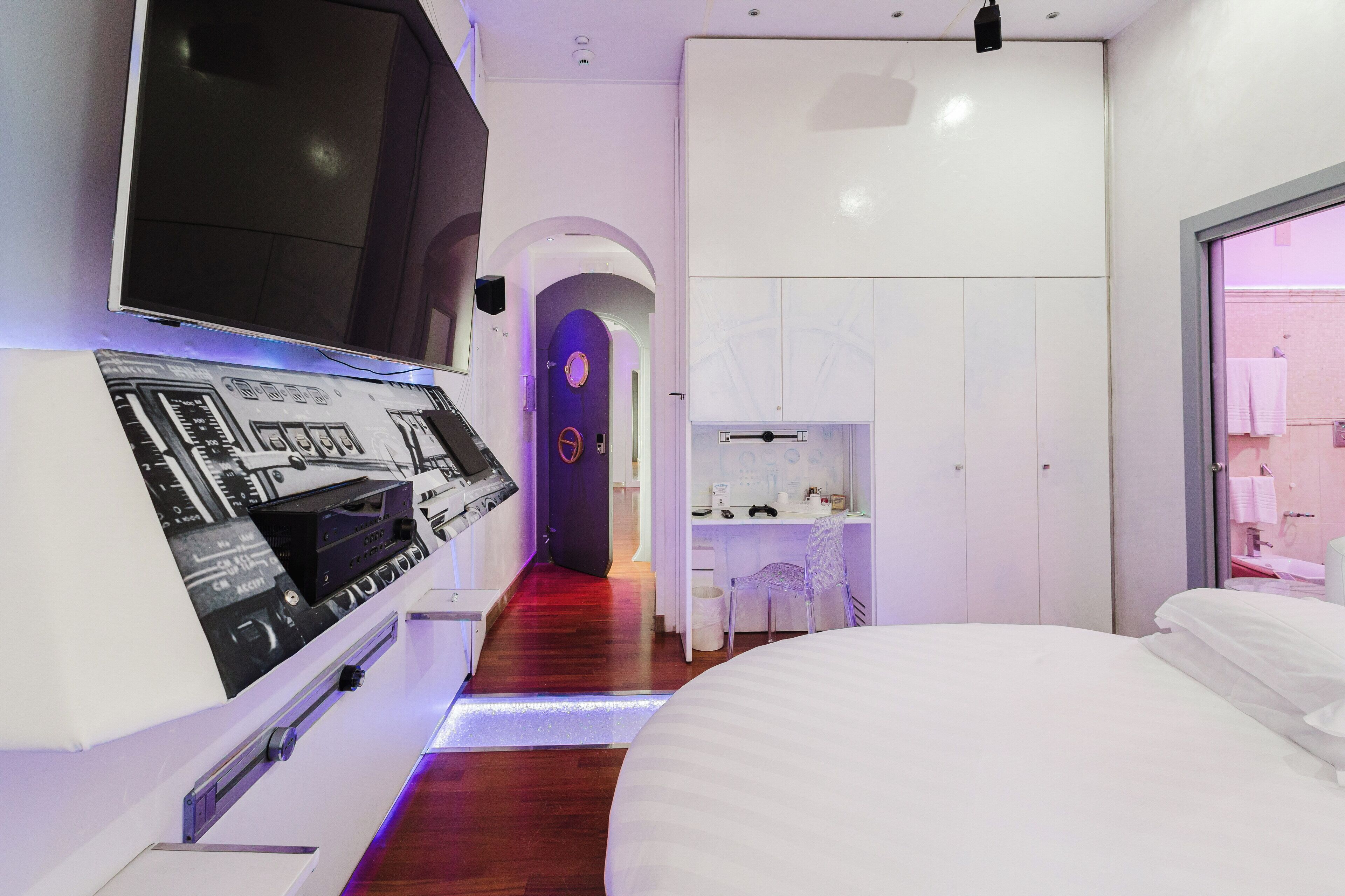 A modern hotel room with a large bed, a flat-screen TV mounted on the wall, a desk with a chair, and a wardrobe.