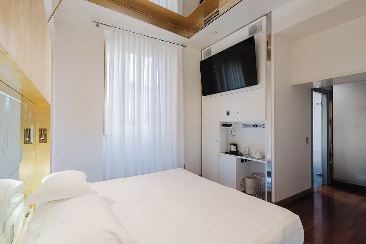 A modern hotel room with a large bed, a flat-screen TV mounted on the wall, a minibar, and a view of the hallway.