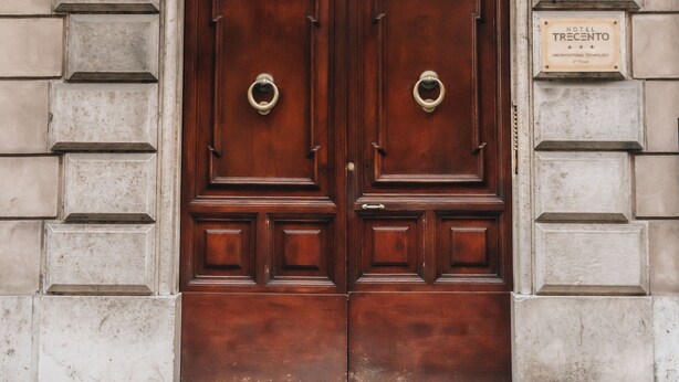 A large, ornate wooden double door with a brass knocker, set within a stone facade with a balcony above. - Hotel Trecento (Rome)