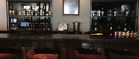 Una zona de bar con varias estanterías llenas de botellas de licores, una máquina de café y taburetes con cojines rojos.