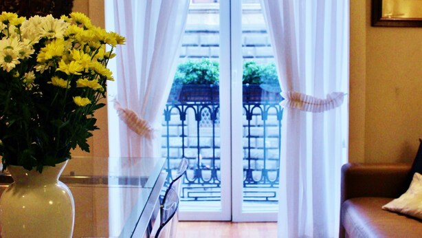 Una habitación con una mesa de vidrio, un jarrón de flores, un sofá de cuero marrón y vistas a través de una puerta con cortinas blancas.