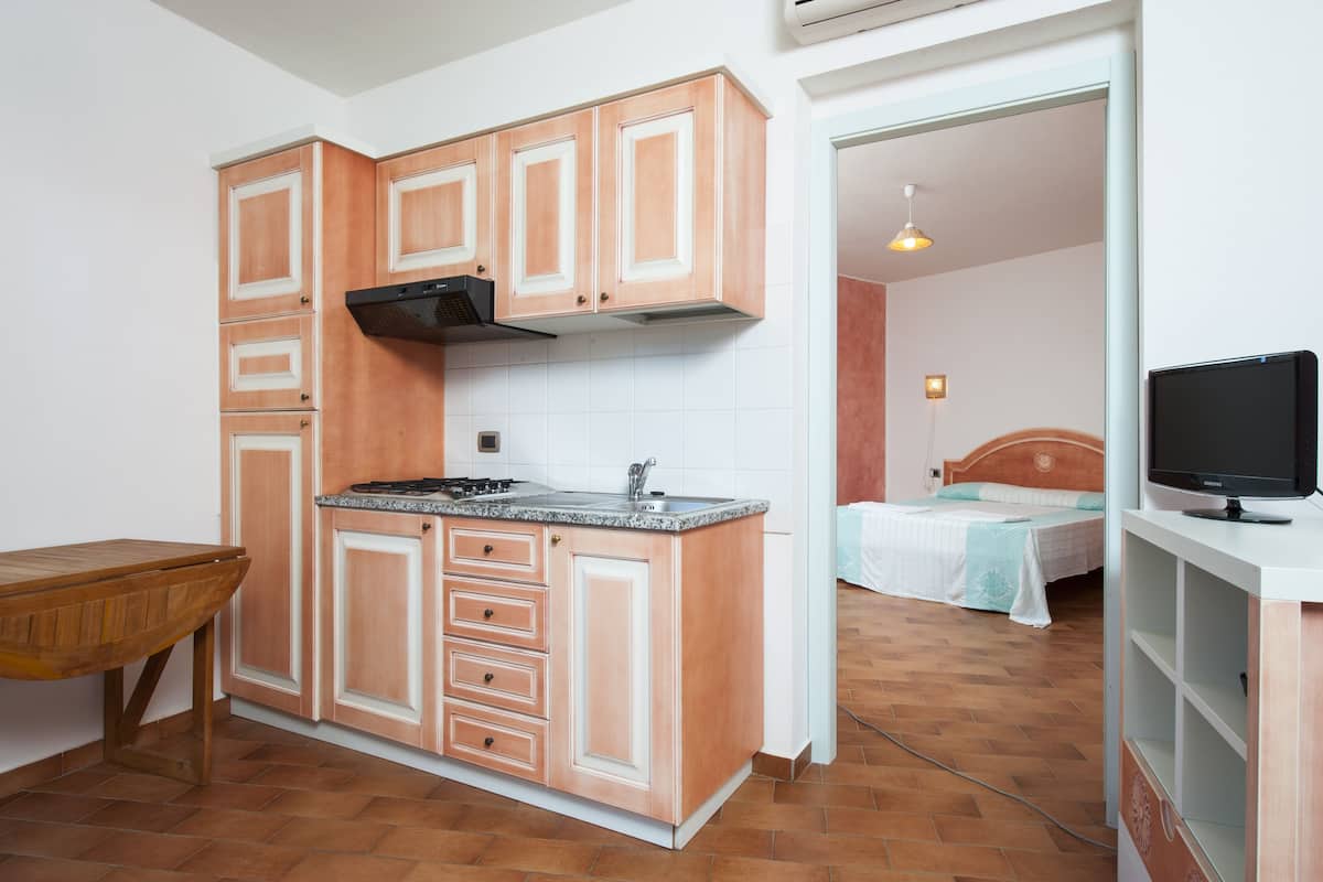 Une cuisine compacte avec des meubles en bois, un plan de travail et une cuisinière. La pièce adjacente contient un lit avec une tête de lit.