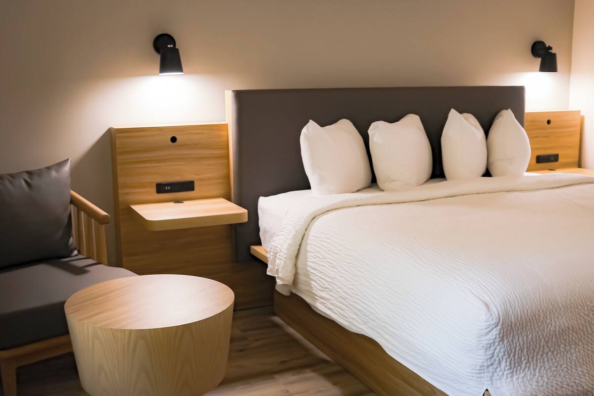 Een moderne slaapkamer met een groot bed, twee nachtkastjes, een rond houten krukje en een kleine bank.