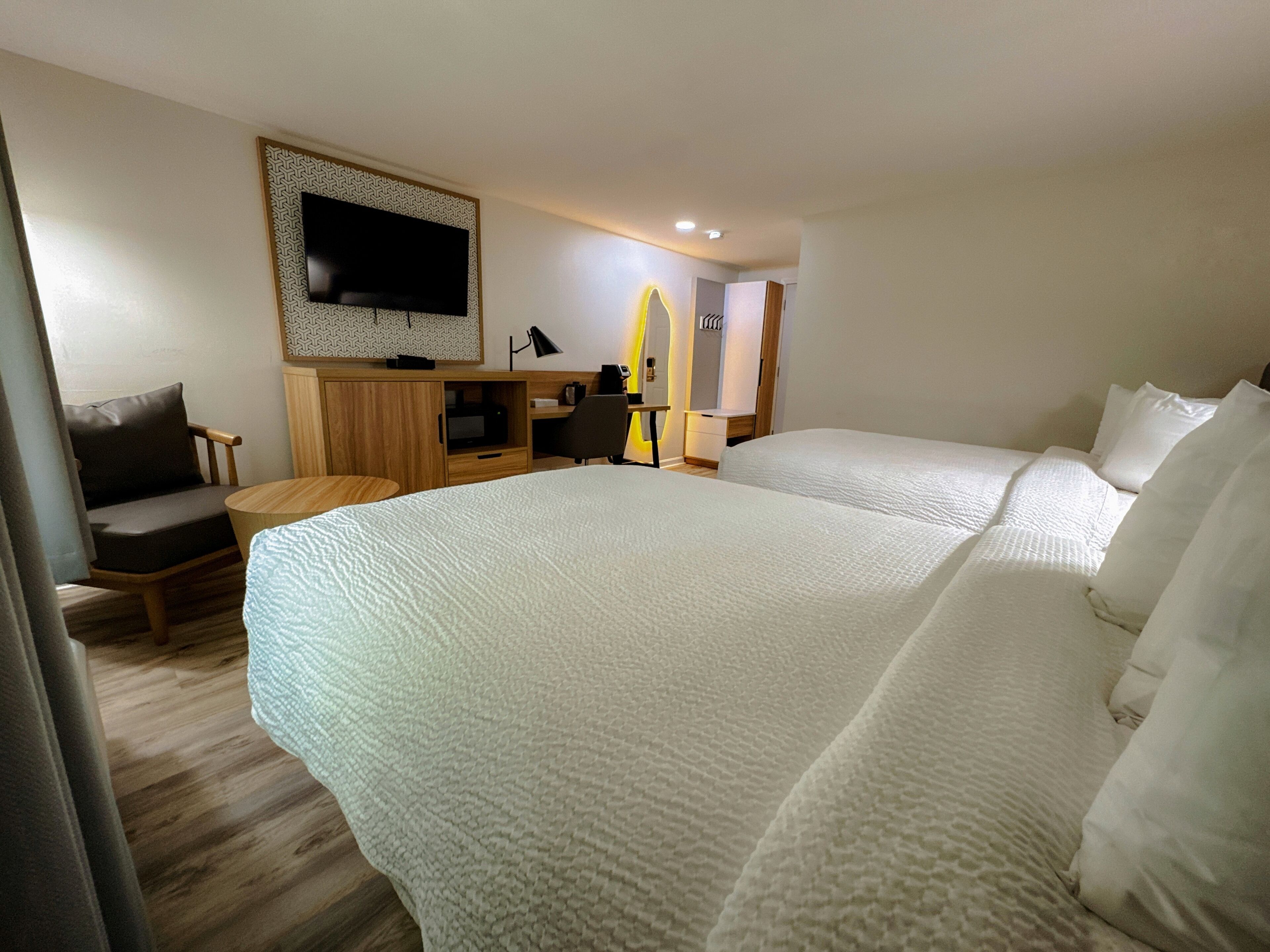 Habitación de hotel con dos camas grandes, un televisor montado en la pared, un escritorio con una silla y una mesita.