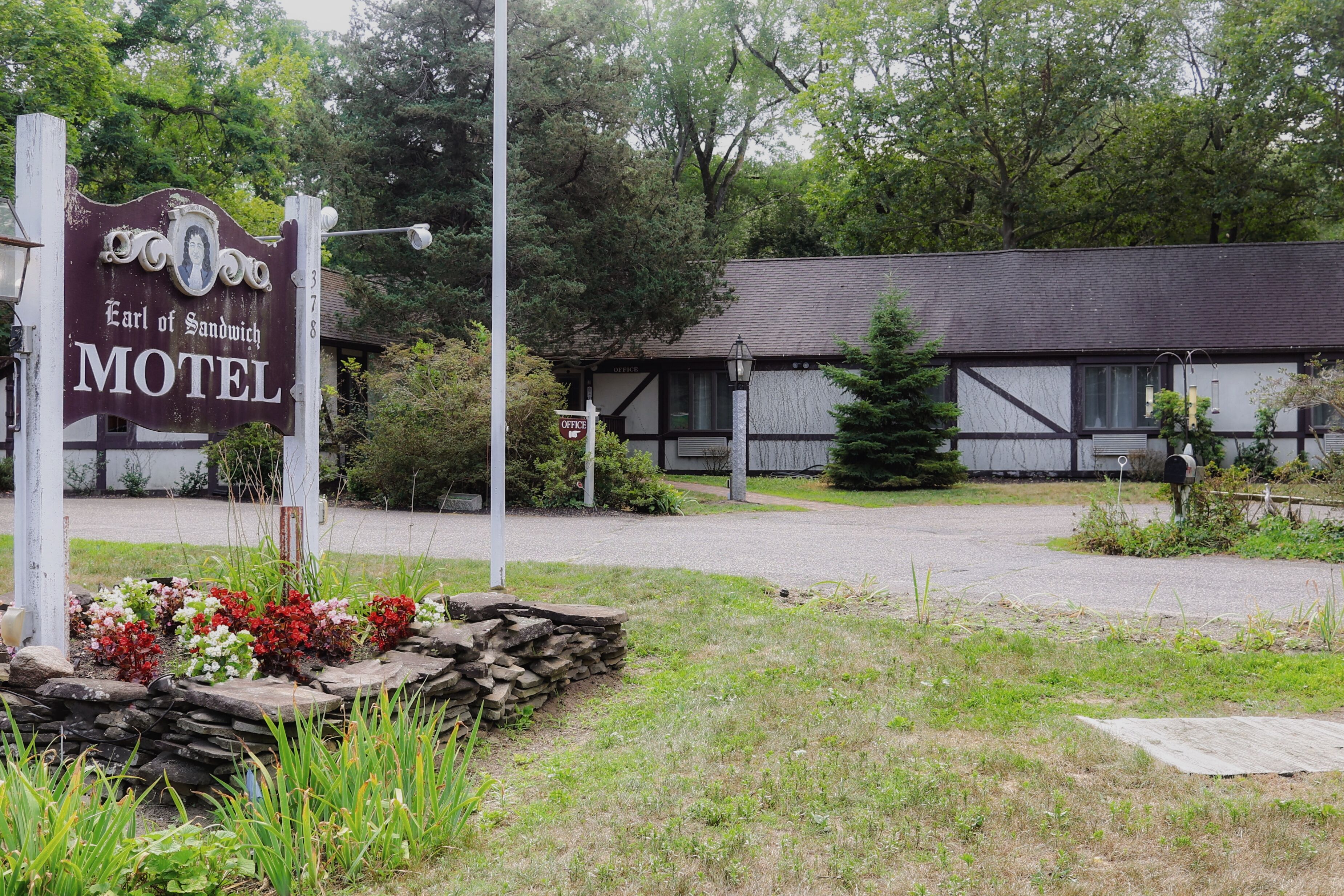 A motel sign with a traditional design, a stone flower bed, and a building with a dark roof and white trim.