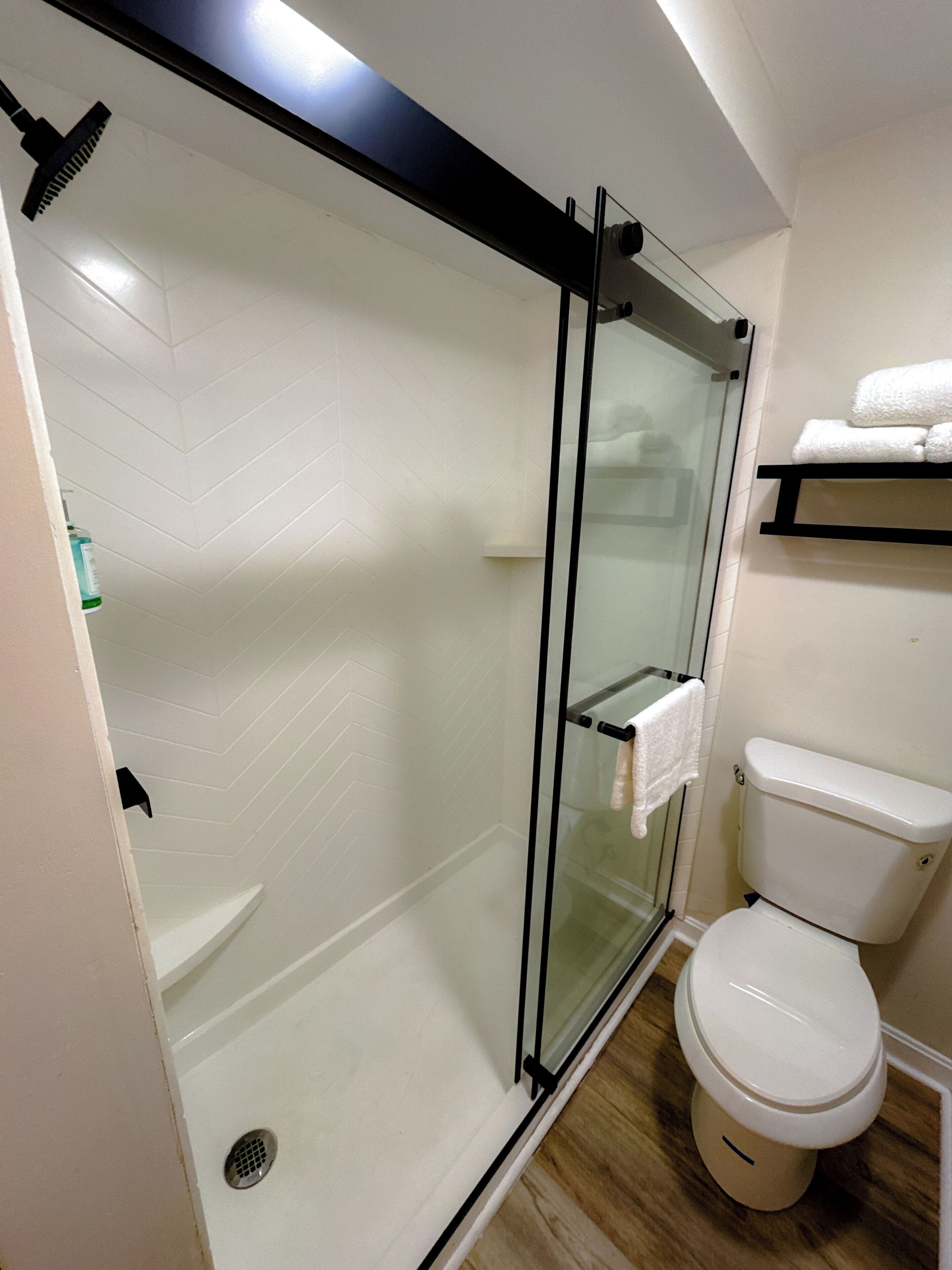 A modern bathroom with a glass-enclosed shower, a white toilet, and a shelf with towels.