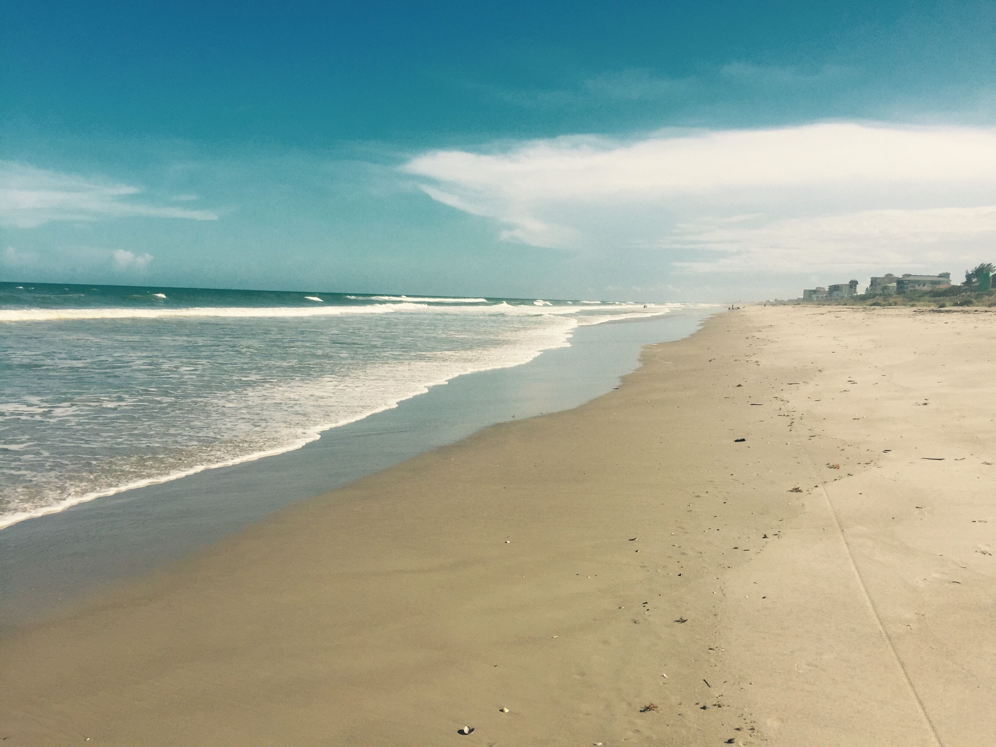 A wide sandy beach with gentle waves, a clear sky, and buildings in the distance.